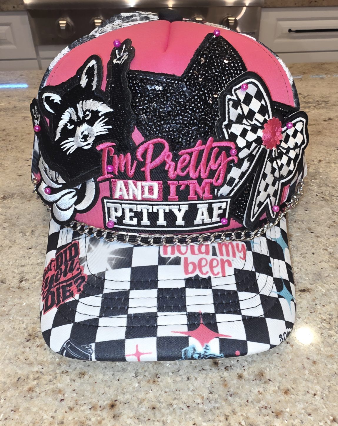 Pink and black checkered trucker hat with a raccoon patch, black sequin cat, checkered bow, 'I'm Pretty and I'm Petty AF' embroidered lettering, and a silver chain across the brim