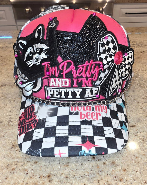 Pink and black checkered trucker hat with a raccoon patch, black sequin cat, checkered bow, 'I'm Pretty and I'm Petty AF' embroidered lettering, and a silver chain across the brim